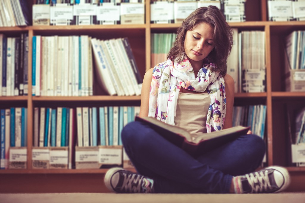 Full length of a female student sitting against bookshelf and re